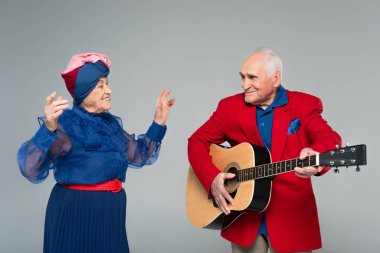Kırmızı ceketli, gülümseyen yaşlı bir adam akustik gitar çalıyor. Gri elbiseli, dans eden kadının yanında.