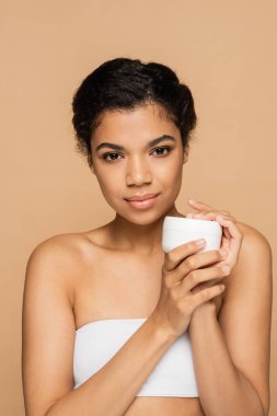 young african american woman with bare shoulders holding container with cosmetic cream isolated on beige  clipart
