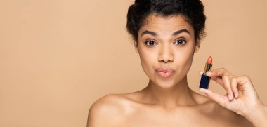 Afro-Amerikan kadın dudaklarını büzüyor ve elinde bej renkli pankartta izole edilmiş ruj tüpü tutuyor.