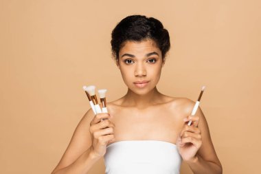 brunette african american woman with bare shoulders holding cosmetic brushes isolated on beige  clipart