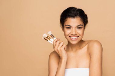 happy young african american woman with bare shoulders holding cosmetic brushes isolated on beige  clipart