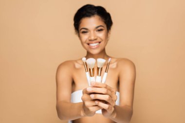 blurred and cheerful african american woman with bare shoulders holding cosmetic brushes isolated on beige  clipart