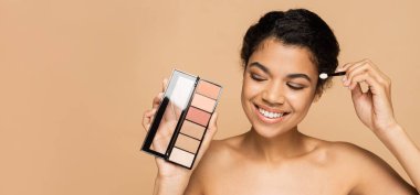 happy african american woman with bare shoulders holding cosmetic brush and blush palette isolated on beige, banner clipart
