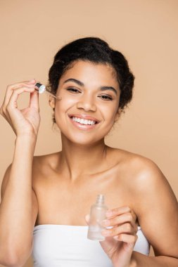 Mutlu bir Afro-Amerikan kadını çıplak omuzlarıyla bej rengi pipetli serum uyguluyor. 