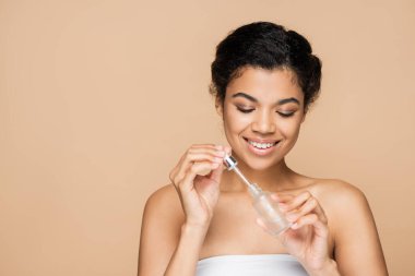 Çıplak omuzlu, elinde serum şişesi ve bej rengi pipetle mutlu bir Afrikalı Amerikalı kadın. 