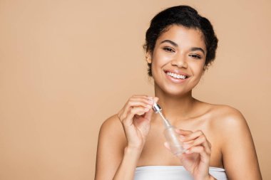 Serum şişesi ve bej üzerinde izole edilmiş pipetle çıplak omuzlu neşeli Afro-Amerikalı kadın. 