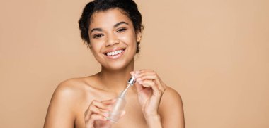 happy african american woman with bare shoulders holding serum bottle and pipette isolated on beige, banner clipart