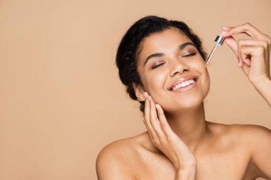 happy african american woman with bare shoulders applying serum isolated on beige  clipart