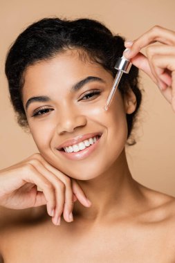 Çıplak omuzlu neşeli Afrikalı Amerikalı kadın bej üzerine serum uyguluyor. 