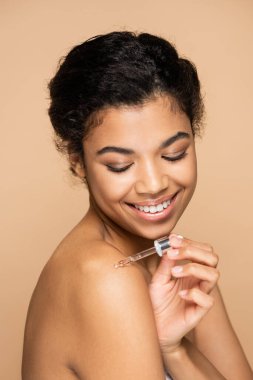 Çıplak omuzlarına serum süren, bej renkte izole edilmiş mutlu bir Afrikalı Amerikalı kadın. 