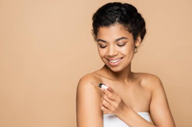 happy african american woman with bare shoulders applying vitamin c serum on bare shoulder isolated on beige  clipart