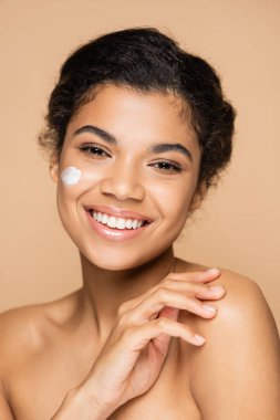 joyful african american woman with face cream on cheek isolated on beige  clipart