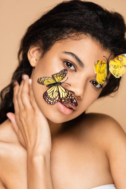 Yüzünde dekoratif kelebekler olan Afrikalı Amerikalı genç bir kadın bej rengi kameraya bakıyor.