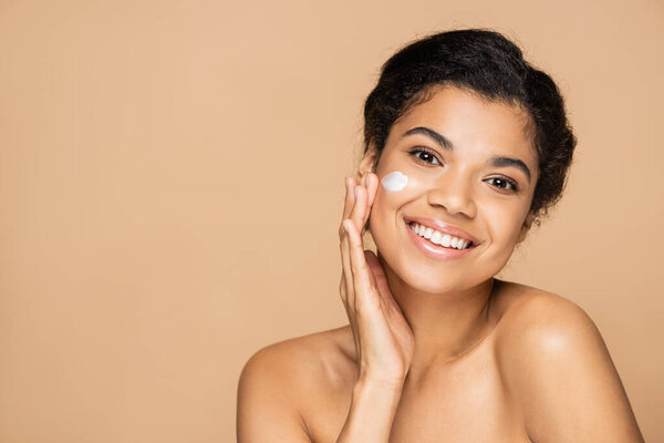 happy african american woman applying face cream on cheek isolated on beige 