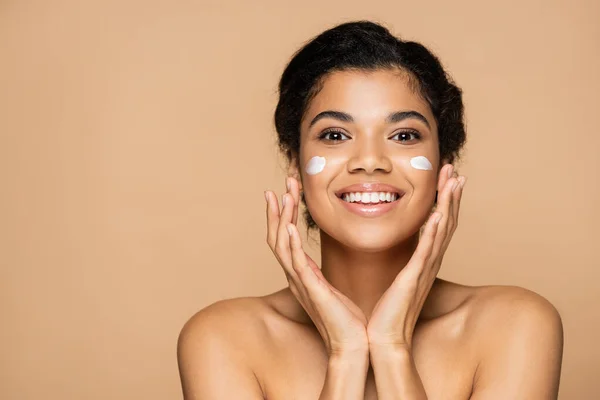 joyful african american woman with face cream on cheeks isolated on beige