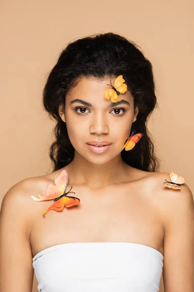 pretty african american woman with orange butterflies on face and bare shoulders looking at camera isolated on beige