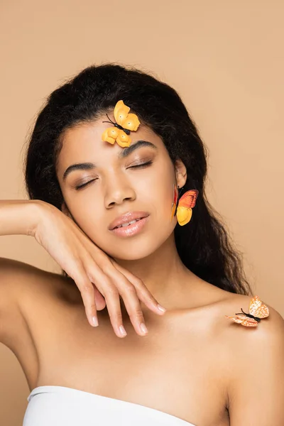 pretty african american woman with closed eyes, orange butterflies on face and bare shoulders isolated on beige