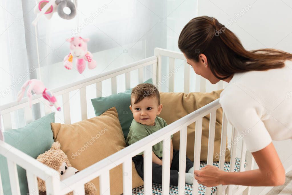 Smiling kid sitting in baby bed near mother 