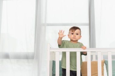 Küçük çocuk bebek yatağında duruyor ve el sallıyor. 