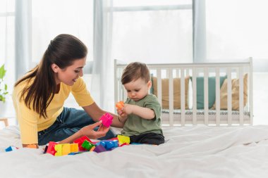 Küçük çocuk yatak odasında gülümseyen annenin yanında yapı taşı oynuyor. 