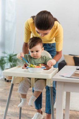 Anne çocuğun yanında duruyor ve yüksek sandalyede yemek yiyor. 
