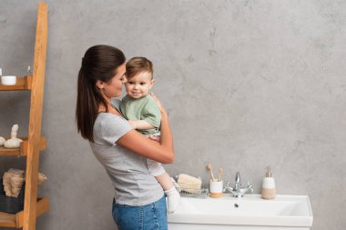 Neşeli anne banyoda lavabonun yanında yürümeye başlayan oğlunu kucaklıyor. 