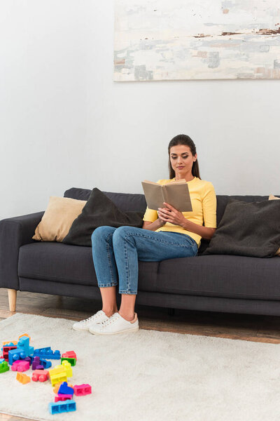 Young woman reading book near building blocks on carpet 