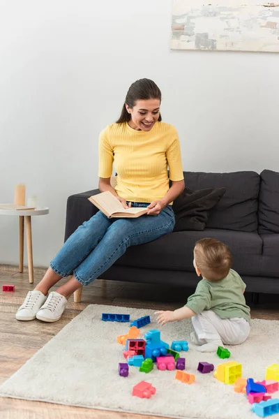 Feliz Madre Sosteniendo Libro Cerca Niño Con Bloques Construcción Alfombra —  Fotos de Stock