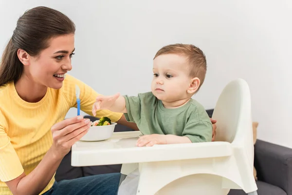 Sorridente Madre Che Tiene Cucchiaio Vicino Bambino Verdure Ciotola Casa — Foto Stock