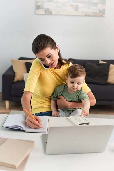 Freelancer Hablando Smartphone Escribiendo Notebook Cerca Niño Pequeño Con Calculadora — Foto de Stock