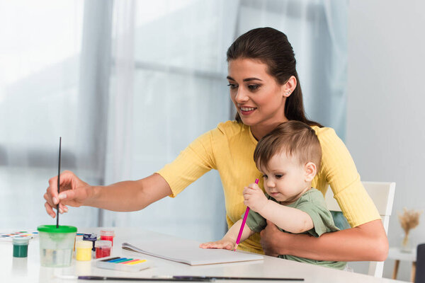 Smiling woman holding paint brush near child with pencil 