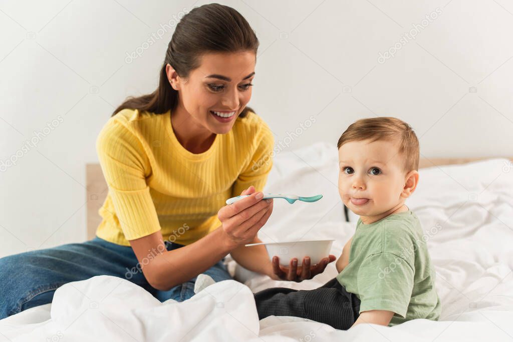 Smiling mother holding spoon near son on bed 