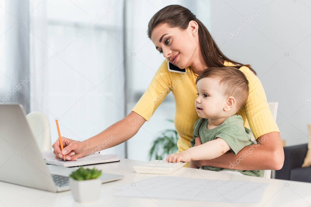 Mother talking on mobile phone while working near toddler son at home 