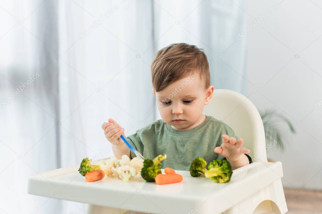 Kid with spoon looking at vegetables on high chair at home 