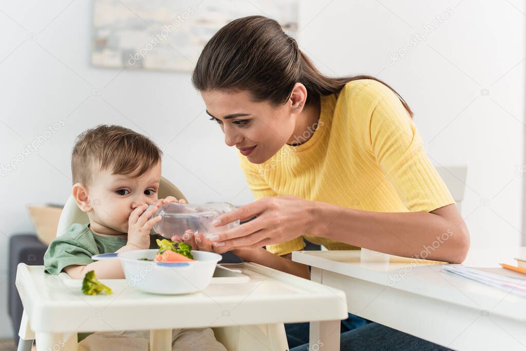 Toddler boy drinking water near mother and food on high chair 