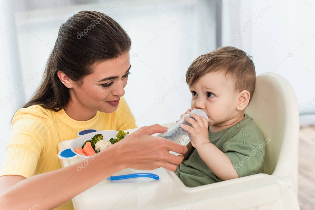 Mother holding baby bottle near son and vegetables on high chair at home 