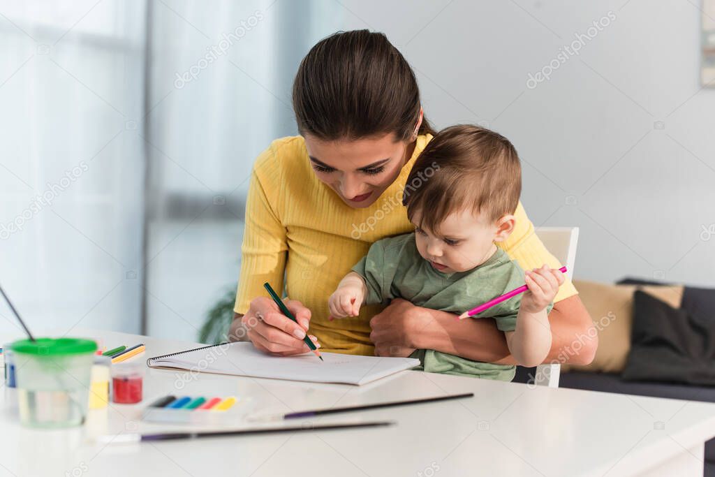 Woman drawing with toddler kid near gouache on blurred foreground 