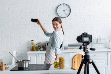 Gülümseyen ev hanımı ocağın yanında selfie çekiyor ve mutfakta bulanık dijital kamera var.