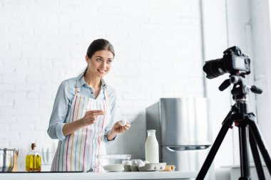 Mutlu mutfak blogcusu bulanık dijital kameraya bakarken tavuk yumurtasını un ile kaseye kırıyor.