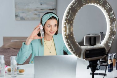 Şık bir kadın dizüstü bilgisayarın yanında su şişesi ve yüzük lambasıyla telefon kulpu gösteriyor.