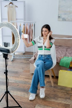 Güzel ve pozitif moda blogcusu kıyafetlerin yanında selfie çekiyor ve daire lambasıyla telefon tutuyor.