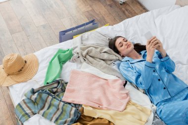 Moda pijamalı gülümseyen bir kadın cep telefonuyla yataktaki çeşitli kıyafetlerin yanında blog yazıyor.