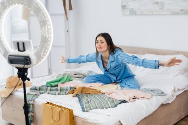 Üzgün moda blogcusu. Kolları açık. Yatak odasında bir sürü kıyafet ve telefon kulübesi var.