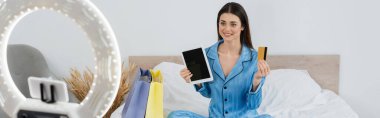 Dijital tableti ve kredi kartı olan moda blogcusu alışveriş çantaları ve telefon tutucusunun yanında gülümsüyor, afiş