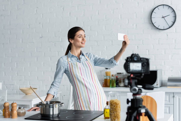 happy culinary blogger taking selfie near blurred digital camera in kitchen