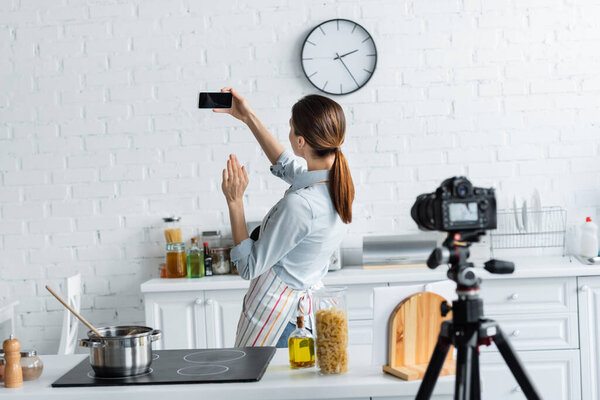 young culinary blogger waving hand near smartphone with blank screen and blurred digital camera in kitchen