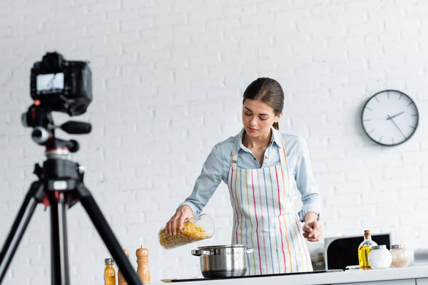 culinary blogger in apron preparing pasta in front of blurred digital camera 