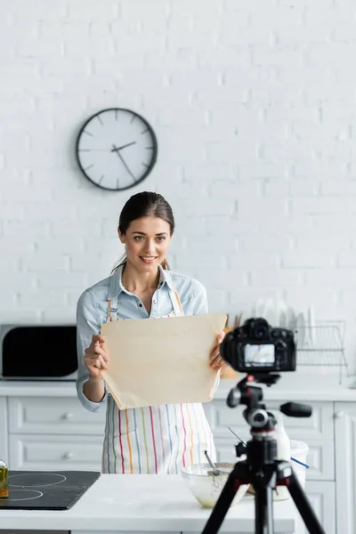 キッチンのぼやけたデジタルカメラでベーキングパーチメントを示す笑顔の料理ブロガー — ストック写真