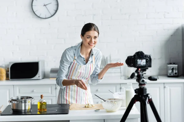 stock image cheerful culinary blogger pointing with hand near dough and blurred digital camera