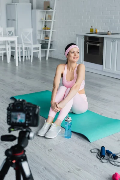 happy sports blogger sitting on fitness mat near blurred digital camera